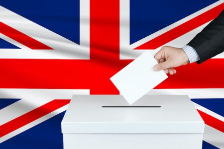Election in United Kingdom. The hand of man putting his vote in the ballot box. Waved United Kingdom flag on background.