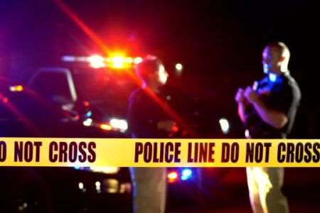 A crime scene at night. Police cars with emergency lights flashing behind yellow cordon tape declaring - police line, do not cross. Two police officers are out of focus talking in the background.