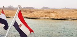 The national flag flutters in the wind against the azure water of the Red Sea and the rocky sandy shore. Close-up. Landscape of the Red Sea. Excursion on a ship to Sharm El Sheikh