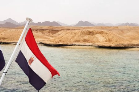 The national flag flutters in the wind against the azure water of the Red Sea and the rocky sandy shore. Close-up. Landscape of the Red Sea. Excursion on a ship to Sharm El Sheikh